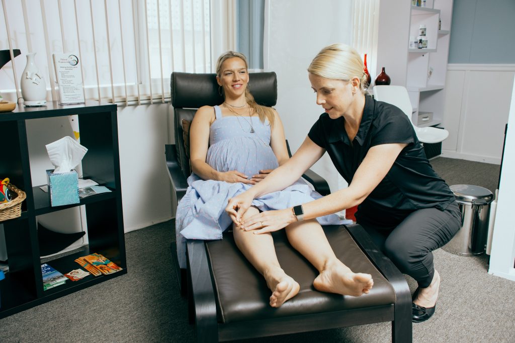 pregnant woman sits in a somewhat reclining chair  with her feet raised while another woman places acupuncture needle at acupuncture point. 