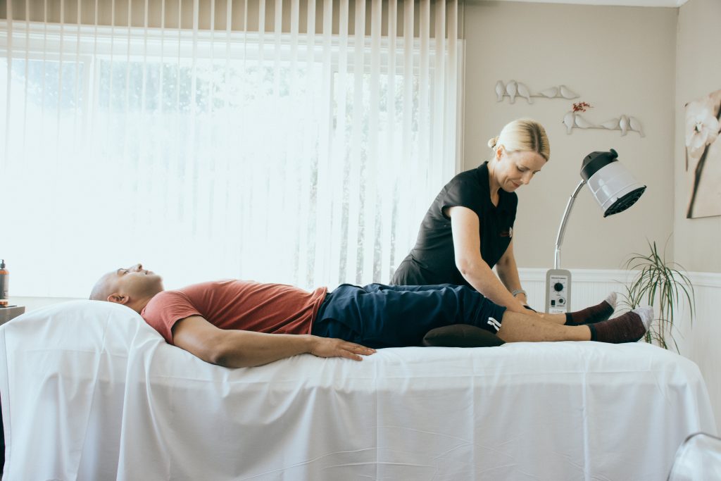 A man lies on a treatment bed with his head and knees supported with a pillow. Tracy is pulling up a pant leg in order to access acupuncture points for treatment. There is sunlight coming through the blinds, the room is bright and welcoming. 